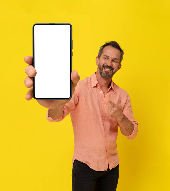 Happy middle aged grey haired fit man holding big smartphone pointing finger at it wearing peach shirt isolated on yellow. Mature muscled man with phone app advertisement. Mock up white screen.  - Φωτογραφία, εικόνα