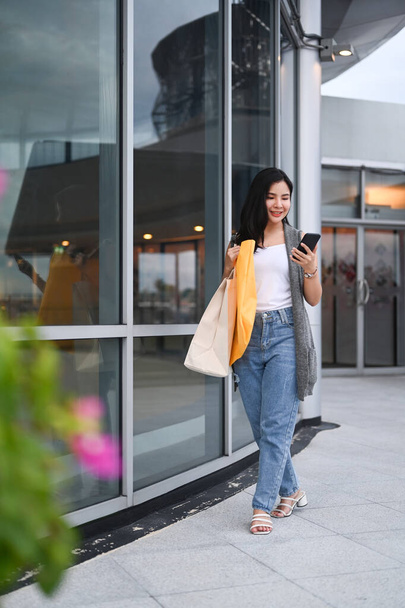 Портрет молодой женщины с сумками с помощью смартфона и прогулкой по городской улице в вечернее время. - Фото, изображение