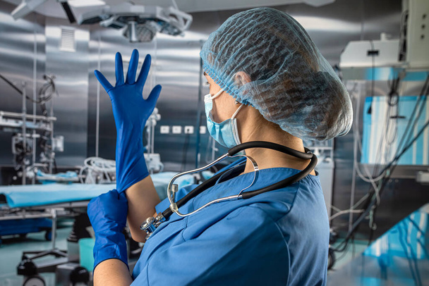 médico o enfermera en uniforme de abrigo azul se pone guantes azules de goma preparación examinar paciente en quirófano - Foto, Imagen