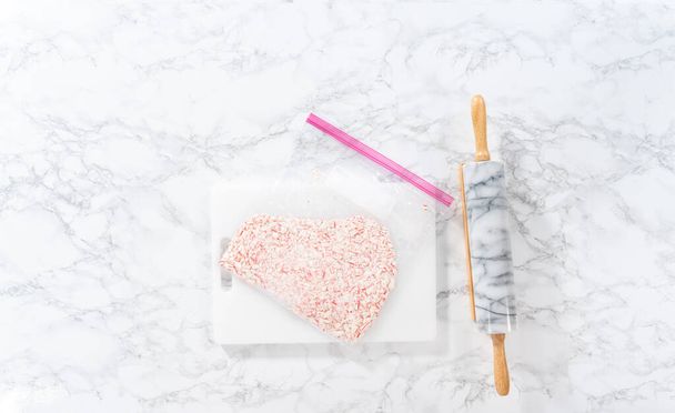 Flat lay. Crushing peppermint candies in a plastic bag with a marble rolling pin on a white cutting board. - Fotoğraf, Görsel