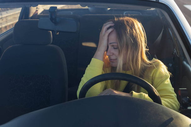 a girl of European appearance with blond hair in a yellow sweater sits behind the wheel of a car, sad, holds her head, does not know what to do, the car broke down on the road. High quality photo - Foto, afbeelding