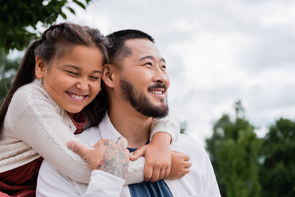 Cheerful asian daughter hugging tattooed father in park  - Zdjęcie, obraz