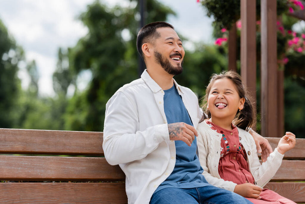 Asian kid sticking out tounge near cheerful dad on bench in park  - Photo, Image