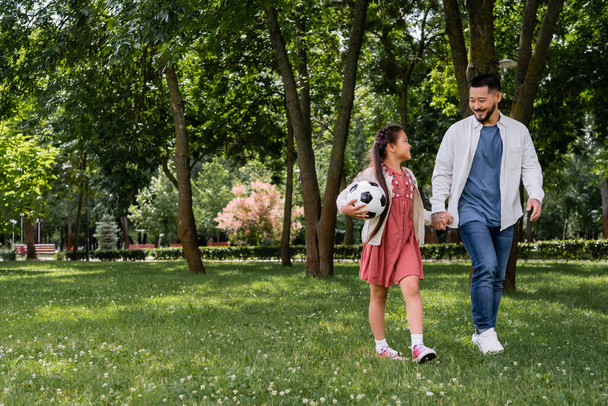 Smiling asian girl holding football and hand of father in park  - Photo, image