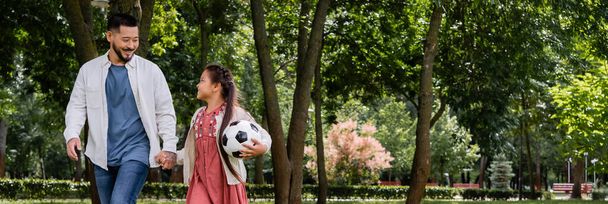 Улыбающийся азиатский папа держит за руку дочь-подростка с футболом в парке, баннер  - Фото, изображение