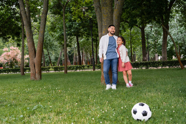 Asian father hugging preteen daughter near tree and soccer ball on grass  - Fotó, kép