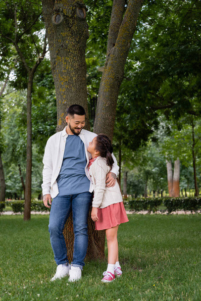 Smiling asian dad hugging and looking at daughter near tree in park  - Fotó, kép