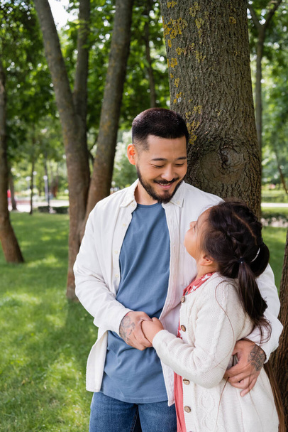 Smiling asian man holding hand and hugging preteen kid near tree in park  - Foto, Imagen