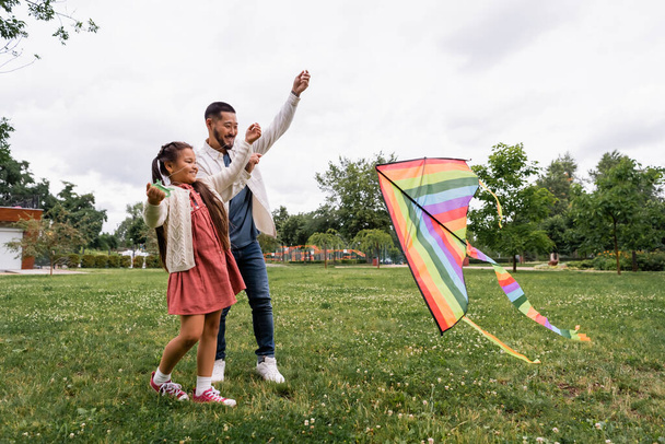Smiling asian parent and kid looking at flying kite on lawn in park  - Foto, imagen