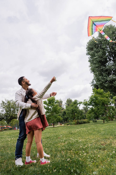 Side view of smiling asian father hugging daughter with flying kite in park  - Foto, Bild