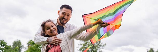 Smiling asian man hugging preteen daughter with flying kite in park, banner  - 写真・画像
