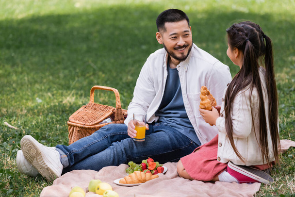 Smiling asian parent holding orange juice near daughter with croissant in park  - Photo, Image