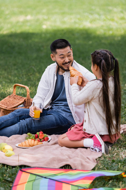 Girl holding croissant near cheerful asian dad with orange juice on blanket in park  - Photo, Image