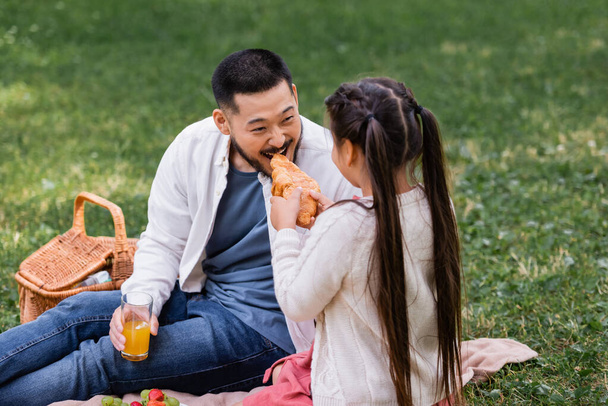 Child holding croissant near asian dad with orange juice in park  - Foto, immagini