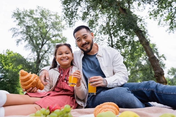 Низкий угол обзора азиатской семьи с апельсиновым соком и круассаном в летнем парке  - Фото, изображение