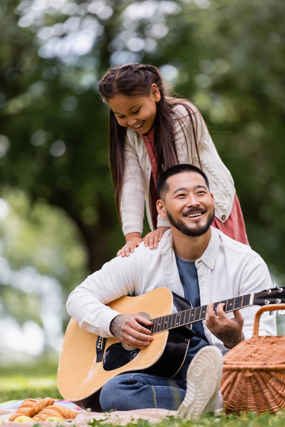 Азійська дитина, яка стоїть поруч з батьком, грає на акустичній гітарі під час пікніка в парку.  - Фото, зображення