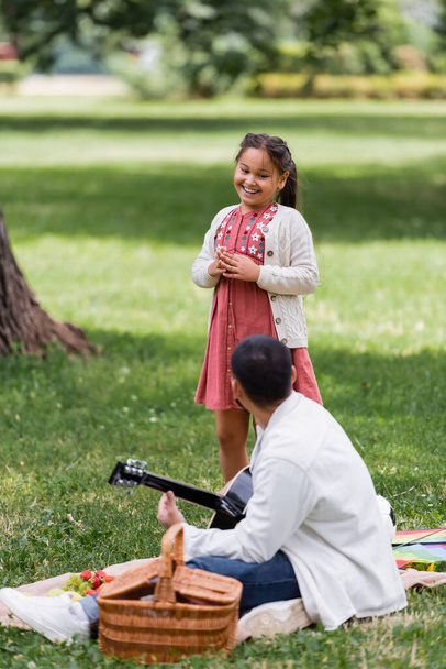 Cheerful asian girl looking at dad playing acoustic guitar during picnic in park  - Zdjęcie, obraz