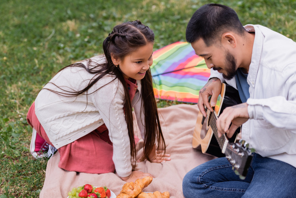 Cheerful asian girl looking at father playing acoustic guitar near fruits and croissants on blanket in park  - Zdjęcie, obraz