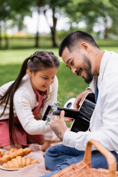 Усміхнений азіатський тато грає на акустичній гітарі біля розмитої дочки та їжі в літньому парку
  - Фото, зображення