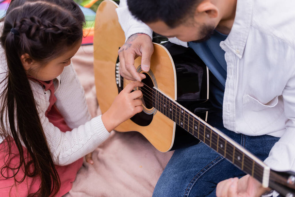 Preteen asian child playing acoustic guitar with father on blanket in park  - Zdjęcie, obraz