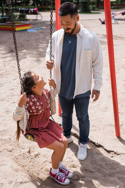 Asian dad looking at daughter on swing in park  - Fotó, kép