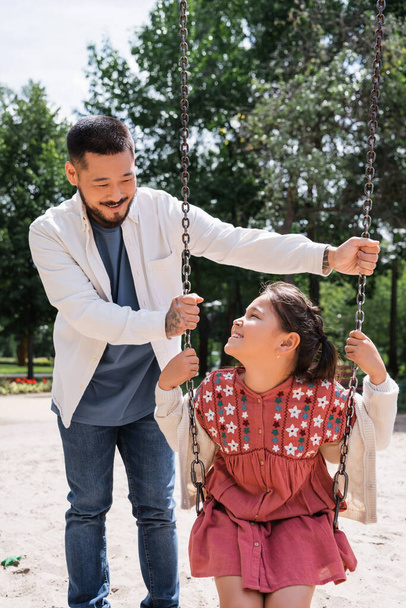 Positive asian girl sitting on swing near tattooed dad in park  - Fotografie, Obrázek