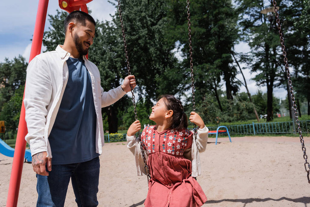 Cheerful asian dad standing near daughter on swing in summer park  - Fotó, kép