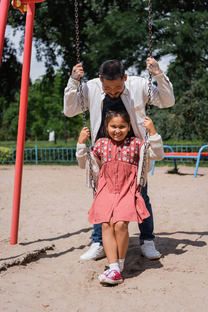Positive preteen girl sitting on swing near asian dad in park  - Фото, изображение