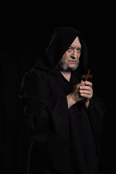 medieval monk in hooded robe holding crucifix and looking at camera isolated on black - Foto, Imagen