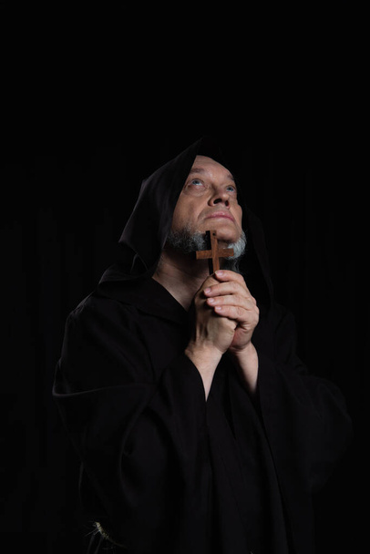 mysterieuze middeleeuwse priester met houten kruis op zoek naar boven geïsoleerd op zwart - Foto, afbeelding