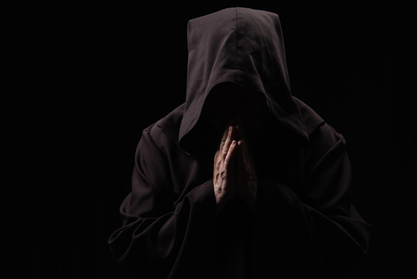 medieval monk with face under dark hood praying isolated on black - Фото, изображение