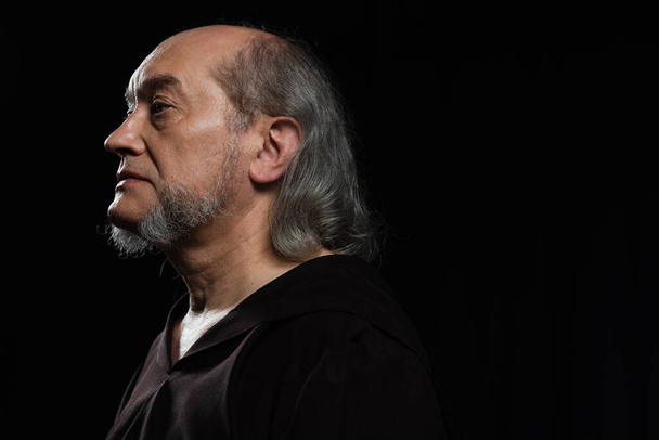portrait de profil de moine sénior aux cheveux gris et à la barbe regardant loin isolé sur noir - Photo, image