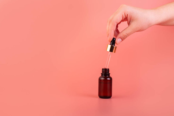 A female hand holds a dropper with a cosmetic serum. Natural cosmetic product for skin purity and hydration. Brown glass bottle with a product for women's face skincare on a pink background - Foto, afbeelding