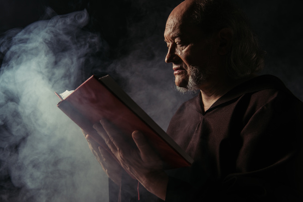 side view of priest in black robe reading holy bible on dark background with smoke - Photo, Image