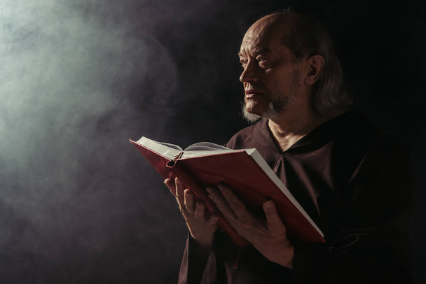 tajemniczy mnich ze świętą biblią odwracający wzrok na czarno z dymem - Zdjęcie, obraz
