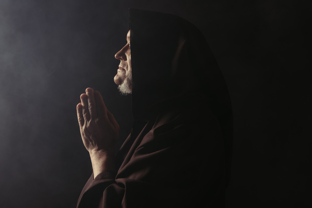 side view of medieval monk in hooded robe praying on black with smoke - Фото, изображение