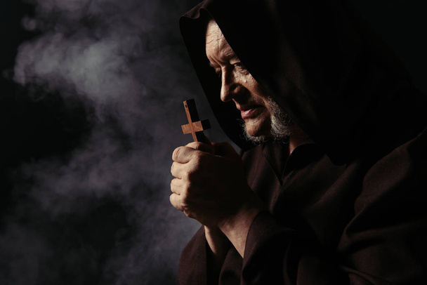 Seitenansicht des Mönchs in schwarzer Kapuzen-Soutane, der mit heiligem Kreuz auf dunklem Hintergrund mit Rauch betet - Foto, Bild