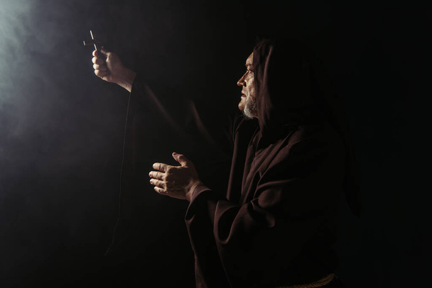 monk in black hooded robe holding holy crucifix in outstretched hand on dark background - Фото, изображение