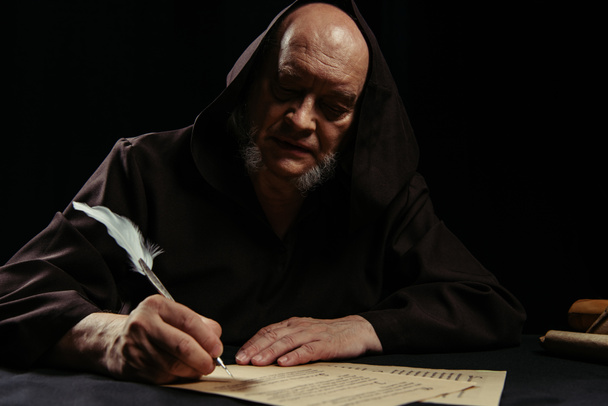 padre em cassock com capuz escuro escrevendo manuscrito à noite isolado em preto - Foto, Imagem
