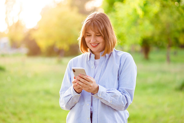 Photo portrait female student in blue shirt keeping cellphone browsing internet walking along summer park - Foto, Imagem