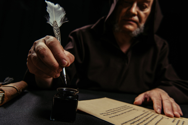 monje borroso sosteniendo pluma pluma cerca de tintero y manuscrito antiguo aislado en negro - Foto, imagen