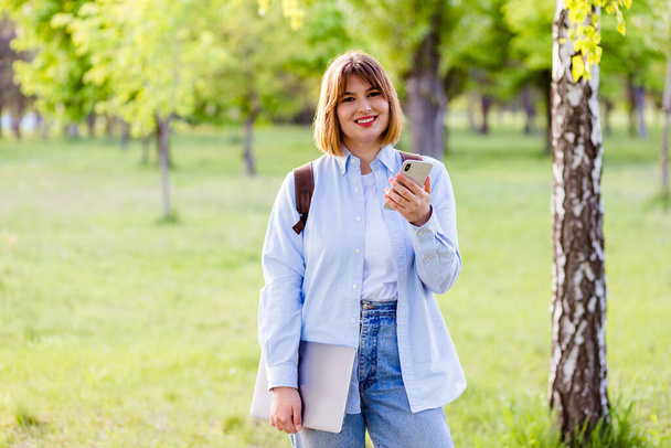 Фотографічний портрет студентки, яка тримає телефон і ноутбук і посміхається на відкритому повітрі в парку
 - Фото, зображення