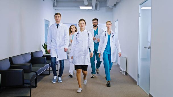 Young and mature team of doctors and nurses walking in front of the camera in the hospital corridor looking straight to the camera concept of medicine and health care. Portrait - Foto, imagen