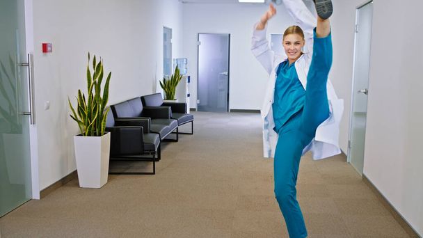 In front of the camera pretty lady doctor in a modern hospital corridor dancing and feeling great she have a cute large smile and great mood. Portrait - Photo, Image
