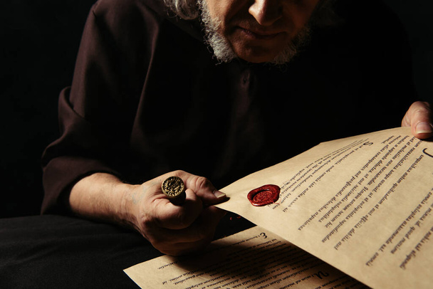 vista cortada de monge sênior segurando pergaminho com selo manuscrito e cera isolado em preto - Foto, Imagem
