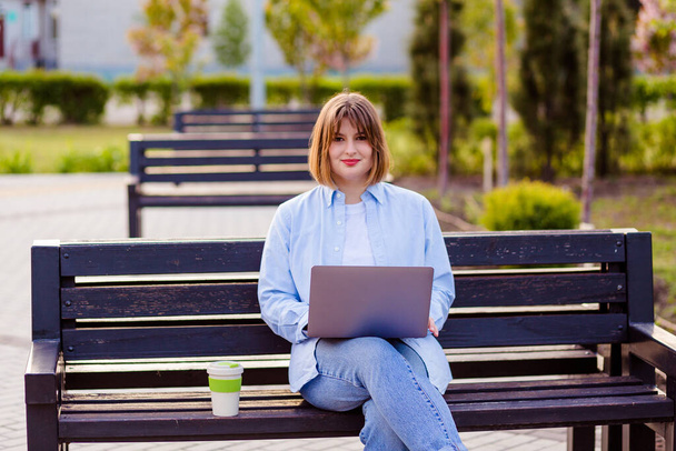 Profile photo of cute lovely young lady sit bench make video call wave hello, greet teacher remote study work online lessons bamboo cup coffee wear white t-shirt jeans park outdoors - Foto, Bild