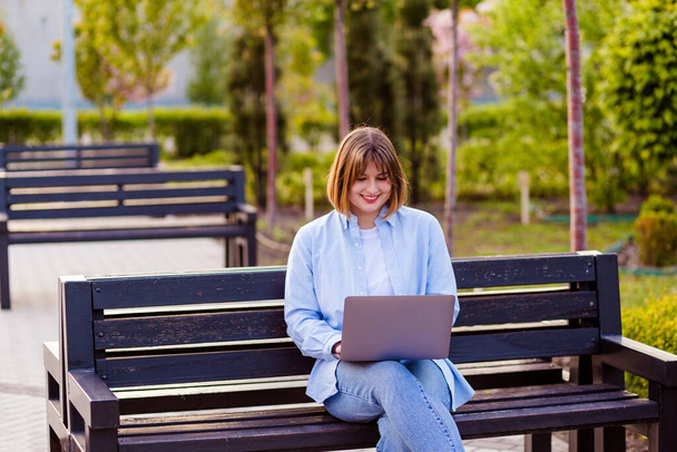 Фото привлекательной студентки сидеть на скамейке в зеленом парке просматривая ноутбук болтая друзья делают домашние задания вдохновение красота природы гармония носить белые футболки парк на открытом воздухе - Фото, изображение