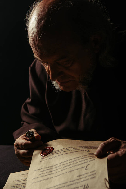 μεσαιωνικός ιερέας κοιτάζοντας κερί σφραγίδα σε αρχαίο χειρόγραφο απομονώνονται σε μαύρο - Φωτογραφία, εικόνα