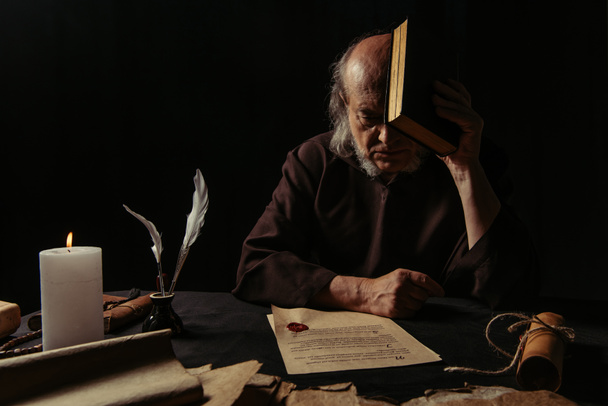 senior priest obscuring face with bible near manuscripts stamped with wax seal  isolated on black - Photo, Image
