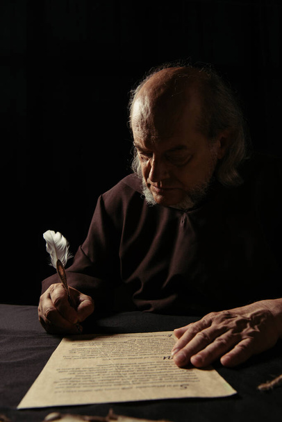 mysterious monk writing chronicle with quill pen in darkness isolated on black - Foto, afbeelding